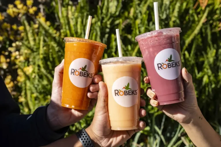 Three hands holding orange, peach, and berry Robeks smoothies outdoors