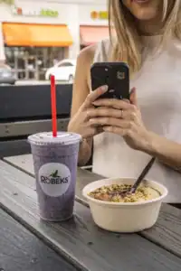 blonde woman takes photo of Robeks smoothie and acai bowl